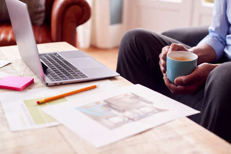 Close Up Of Male Architect Or Interior Designer Working From Home Looking At Designs On Laptop