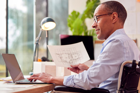Mature Male Architect In Wheelchair Working In Office At Desk Studying Plans For New Buildingの写真素材
