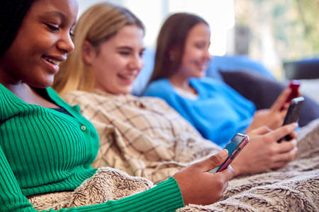 Group Of Multi-Cultural Teenage Girl Friends Snuggled Under Blanket Looking At Mobile Phones At Home
