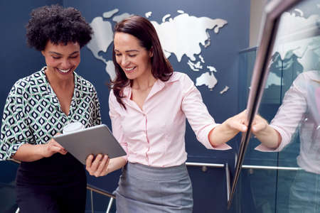 Female Colleagues With Digital Tablet Meeting On Stairs Of Office With World Map In Backgroundの写真素材