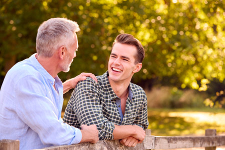 Mature Father With Adult Son Leaning On Fence Walking In Countryside