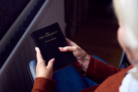 Senior Woman In Wheelchair With Bible Keeping Warm By Radiator At Home In Cost Of Living Crisis