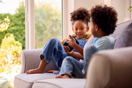 Boy And Girl Playing Handheld Computer Game Sitting On Sofa At Home Together
