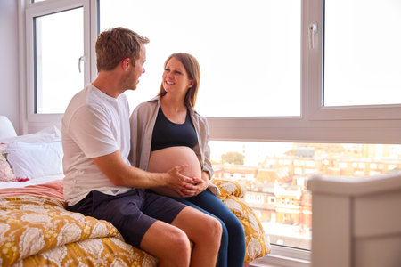 Loving Couple At Home Sitting On Bed With Man Touching Pregnant Woman's Stomach