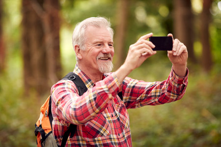 Retired Senior Man Hiking In Woodland Countryside Taking Photo With Mobile Phoneの写真素材