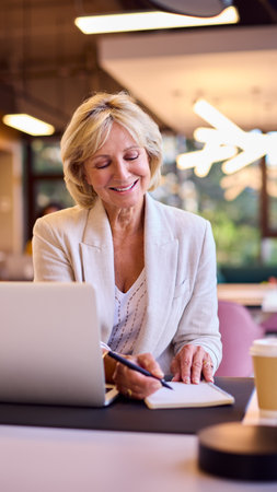 Smiling Mature Businesswoman Working On Laptop At Desk In Office Making Notesの写真素材