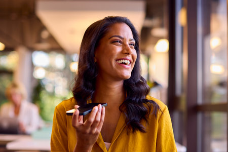 Mature Businesswoman Working At Desk In Office Talking Into Mic Of Mobile Phoneの写真素材