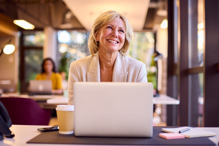 Mature Businesswoman Working On Laptop At Desk In Office With Takeaway Drinkの写真素材