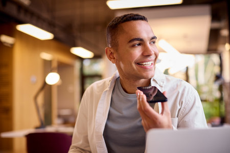 Young Businessman Working On Laptop At Desk In Office Talking Into Mic Of Mobile Phone