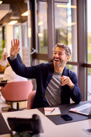 Mature Businessman Working On Laptop At Desk In Office Waving To Colleagueの写真素材