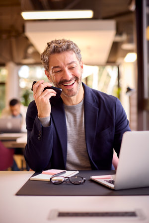 Mature Businessman Working On Laptop At Desk In Office Talking Into Mic Of Mobile Phoneの写真素材