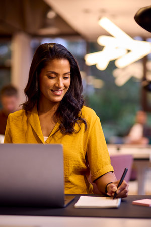 Mature Businesswoman Working On Laptop At Desk In Office Writing In Notebook