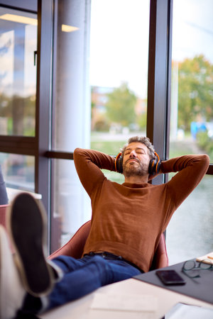 Mature Businessman With Feet On Desk In Office Listening To Music On Wireless Headphonesの写真素材