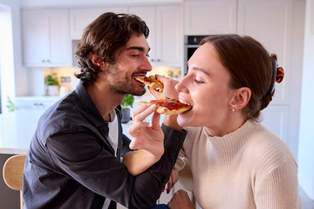 Couple In Kitchen At Home Linking Arms To Eat Homemade Pizza Sitting At Counterの写真素材