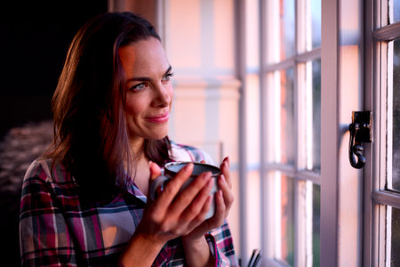 Woman At Home With Hot Drink Standing Looking Out Of Window With Evening Light