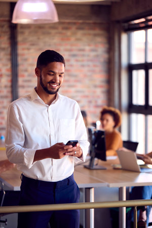 Smiling Businessman Standing In Busy Office Messaging On Mobile Phoneの写真素材