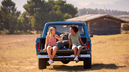 Two Women Riding In Back Of Pick Up Truck As Friends Arrive At Countryside Cabin On Road Trip