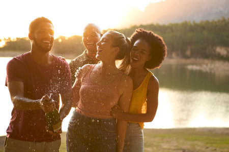 Group Of Friends Celebrating Outdoors By Lake Opening And Spraying Bottle Of Champagneの写真素材
