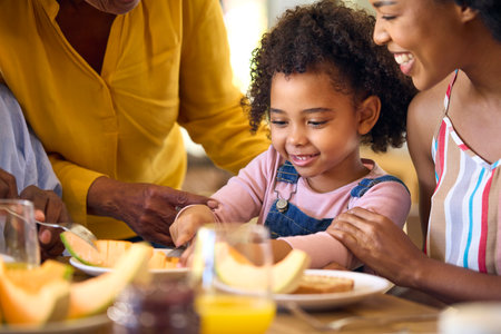 Family Shot With Grandparents Parents And Granddaughter Eating Breakfast Around Table At Homeの写真素材