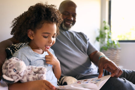 Grandfather Sitting On Sofa At Home Reading Book With Granddaughter