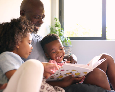 Grandfather Sitting On Sofa At Home Reading Book With Grandchildrenの写真素材