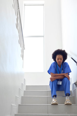 Stressed Female Doctor Or Nurse Wearing Scrubs Sitting On Stairs In Hospitalの写真素材