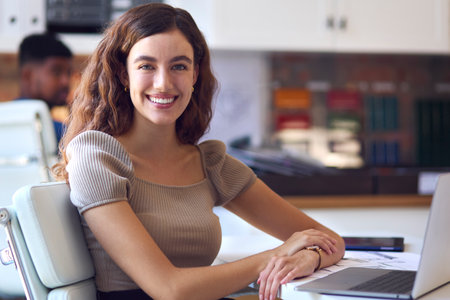Portrait Of Young Smiling Businesswoman Working On Laptop At Desk In Officeの写真素材