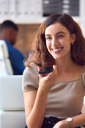 Young Smiling Businesswoman Working At Desk In Office Talking Into Mic Of Mobile Phone