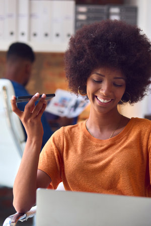 Young Smiling Businesswoman Working At Desk In Office Talking Into Mic Of Mobile Phone