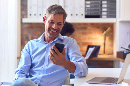 Mature Businessman Working At Desk In Office Messaging On Mobile Phoneの写真素材