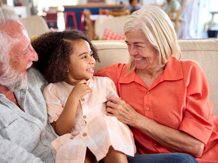 Granddaughter Cuddling With Loving Grandparents On Sofa At Home With Parents In Background
