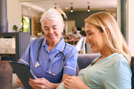 Senior Midwife With Digital Tablet Visiting Pregnant Woman At Home With Family In Backgroundの写真素材