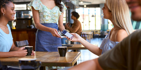 Woman In Coffee Shop Meeting Friend Paying Bill With Contactless Mobile Phone Paymentの写真素材