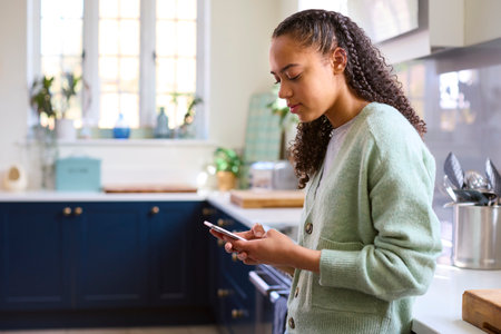 Teenage Girl At Home In Kitchen Connecting With Friends On Social Media Using Mobile Phoneの写真素材