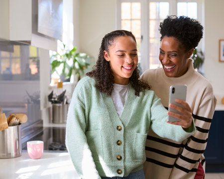 Mother With Teenage Daughter At Home In Kitchen Posing For Selfie On Mobile Phoneの写真素材