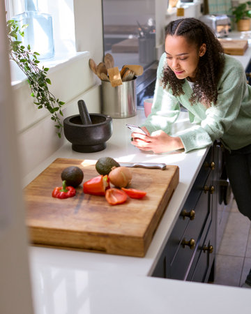 Teenage Girl At Home In Kitchen With Ingredients Looking At Recipe On Mobile Phone