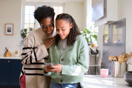 Mother Talking With Teenage Daughter At Home Looking At Social Media On Mobile Phone Together