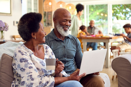 Senior Couple At Home Looking At Laptop Together With Multi-Generation Family In Background