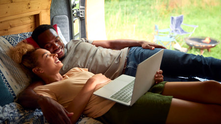 Couple Enjoying Camping In Countryside Lying Inside RV Streaming Movie On Laptop Together