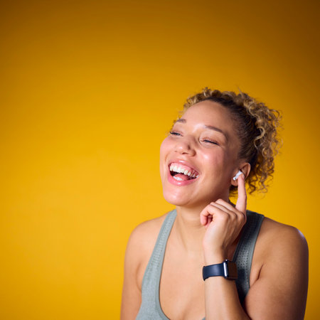 Studio Shot Of Woman In Gym Fitness Clothing Streaming To Wireless Ear Buds On Yellow Background