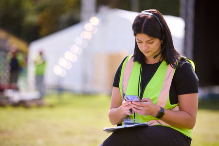 Female Production Worker With Mobile Phone Setting Up Outdoor Stage For Music Festival Or Concert