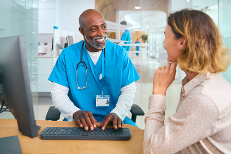 Mature Male Doctor Wearing Scrubs At Hospital Appointment With Female Patient Looking At Computerの写真素材