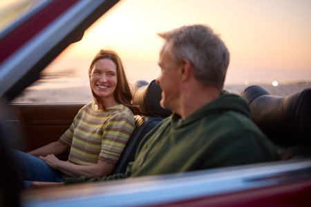 Retired Senior Couple On Vacation Sitting In Classic Sports Car At Beach Watching Sunrise