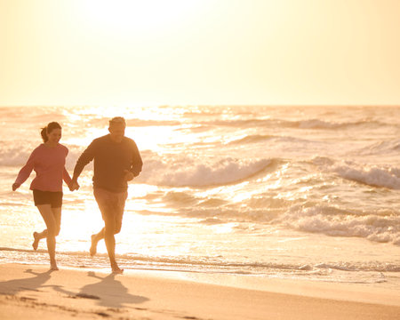 Loving Retired Senior Couple On Vacation Running Along Beach Shoreline Holding Hands At Sunriseの写真素材