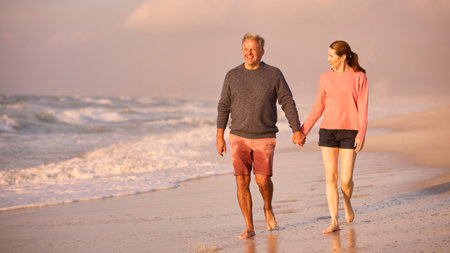 Loving Retired Senior Couple On Vacation Walking Along Beach Shoreline Holding Hands At Sunriseの写真素材