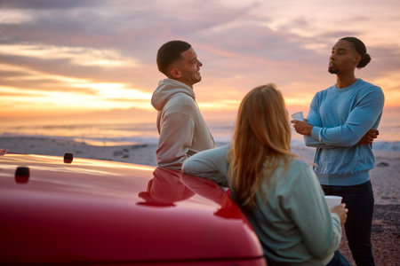 Group Of Friends Standing Chatting By Car With Hot Drink At Beach Watching Sunrise Togetherの写真素材