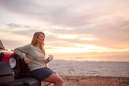 Young Woman Standing By Car With Hot Drink At Beach Watching Sunrise
