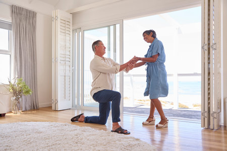 Mature Couple With Man Proposing Marriage Kneeling And Giving Woman Ring In Beach Front Propertyの写真素材