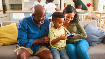 Grandfather With Mother And Grandson Play Handheld Computer Game At Home With Family In Background