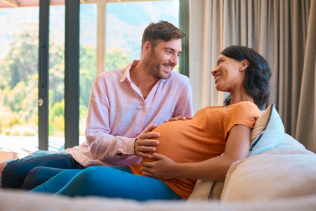Loving Multi-Racial Couple With Pregnant Woman On Sofa And Man Touching Woman's Stomach At Home